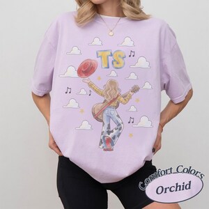 May include: A light purple t-shirt featuring a graphic illustration of a person with long hair wearing a yellow plaid shirt and cow-print bell-bottom pants, holding a guitar and a red hat. The design includes white clouds, yellow stars, and the text TS.