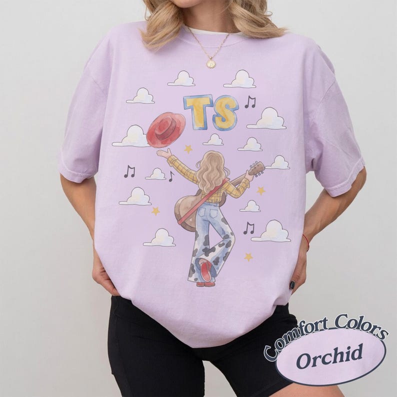 May include: A light purple t-shirt featuring a graphic illustration of a person with long hair wearing a yellow plaid shirt and cow-print bell-bottom pants, holding a guitar and a red hat. The design includes white clouds, yellow stars, and the text TS.