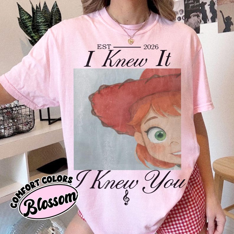 May include: A light pink t-shirt featuring a graphic print of a cartoon character with red hair and a red hat. The text above the graphic reads EST 2026 I Knew It, and the text below reads I Knew You with a small musical note symbol.