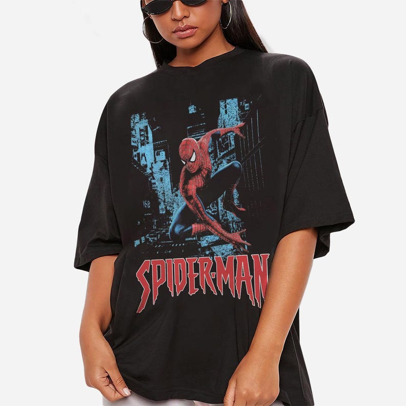 May include: Black t-shirt featuring a graphic of Spider-Man in a city setting. The superhero is depicted in red and blue, with the word "SPIDERMAN" in red block letters below the image. The shirt has short sleeves.