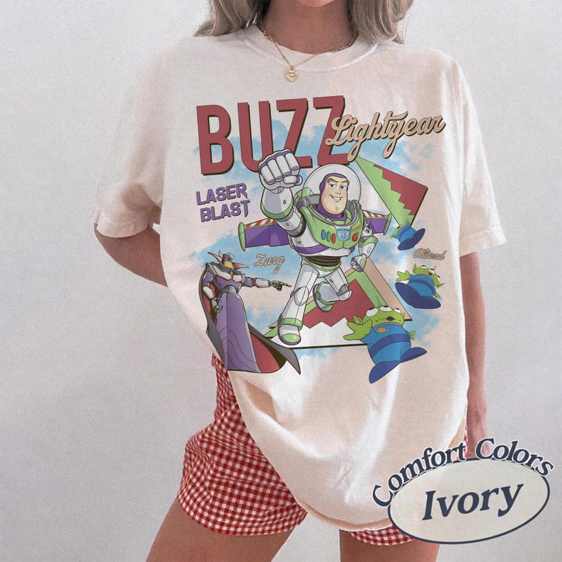 May include: Ivory t-shirt with a graphic of Buzz Lightyear from Toy Story. The design includes the words "Buzz Lightyear" and "Laser Blast," along with images of Zurg and the aliens. The shirt is a Comfort Colors brand.