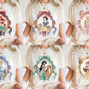 May include: Cream-colored t-shirts featuring watercolor illustrations of Disney princesses within ornate frames. Each shirt showcases a different princess, including Ariel, Snow White, Aurora, Cinderella, Jasmine, and Belle, with accompanying characters and decorative bows.