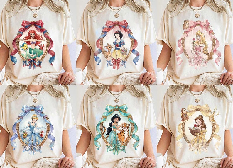 May include: Cream-colored t-shirts featuring watercolor illustrations of Disney princesses within ornate frames. Each shirt showcases a different princess, including Ariel, Snow White, Aurora, Cinderella, Jasmine, and Belle, with accompanying characters and decorative bows.