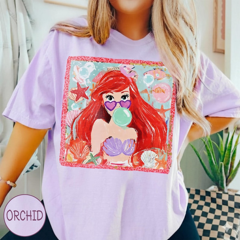 May include: A light purple t-shirt featuring a colorful graphic of a cartoon mermaid with red hair, heart-shaped sunglasses, and a bubble. The graphic is framed with a pink border and surrounded by seashells and starfish. The word "ORCHID" is printed on the bottom left.