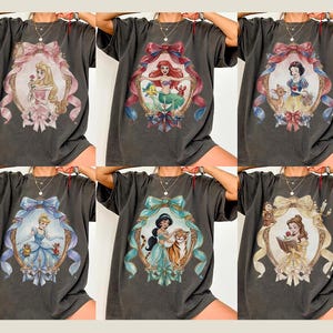 May include: Dark gray t-shirts featuring various Disney princess portraits within ornate frames. Each shirt showcases a different princess, including Aurora, Ariel, Snow White, Cinderella, Jasmine, and Belle, with detailed illustrations and colorful accents.