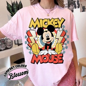 May include: A light pink t-shirt featuring a graphic of Mickey Mouse. The design includes the words "MICKEY MOUSE" in yellow and red, with Mickey Mouse in the center. The shirt also has the words "COMFORT COLORS" and "Blossom" printed on it.