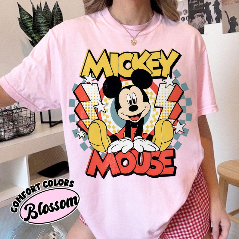 May include: A light pink t-shirt featuring a graphic of Mickey Mouse. The design includes the words "MICKEY MOUSE" in yellow and red, with Mickey Mouse in the center. The shirt also has the words "COMFORT COLORS" and "Blossom" printed on it.