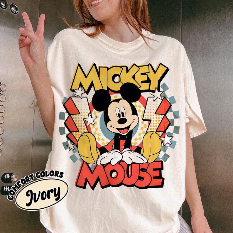May include: Ivory-colored t-shirt featuring a graphic of Mickey Mouse. The design includes the words "Mickey Mouse" in yellow and red, with Mickey in the center. The shirt has a vintage, distressed look, with a "Comfort Colors" label.