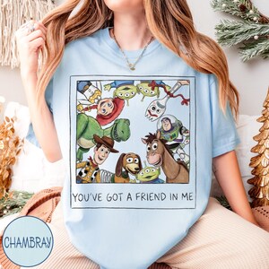 May include: Light blue t-shirt featuring a rectangular graphic with cartoon characters from the movie Toy Story. The graphic includes Woody, Buzz Lightyear, Rex, and the Little Green Men. The text "YOU'VE GOT A FRIEND IN ME" is printed below the graphic.
