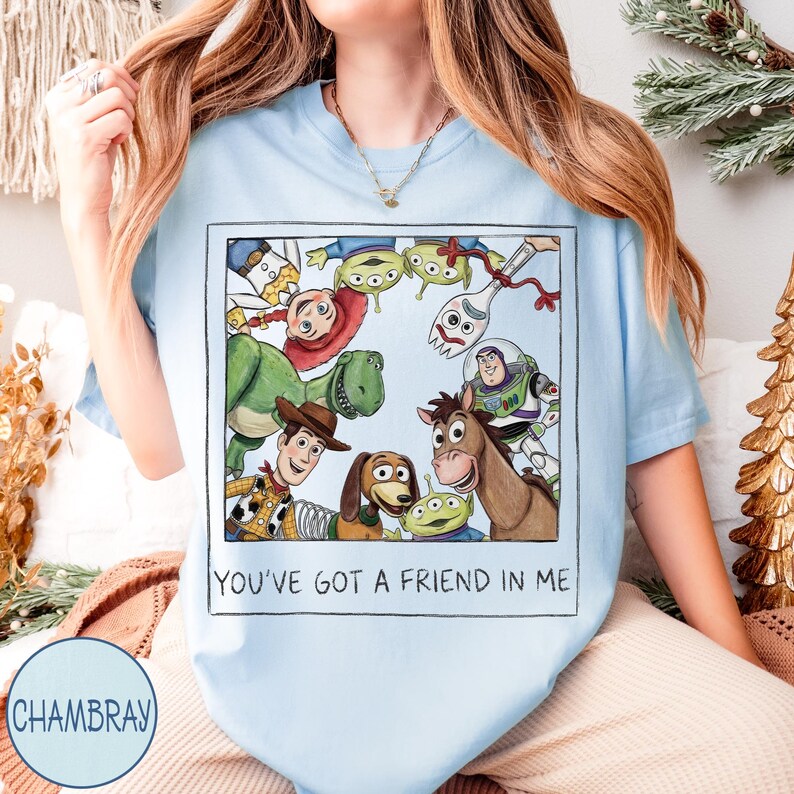 May include: Light blue t-shirt featuring a rectangular graphic with cartoon characters from the movie Toy Story. The graphic includes Woody, Buzz Lightyear, Rex, and the Little Green Men. The text "YOU'VE GOT A FRIEND IN ME" is printed below the graphic.