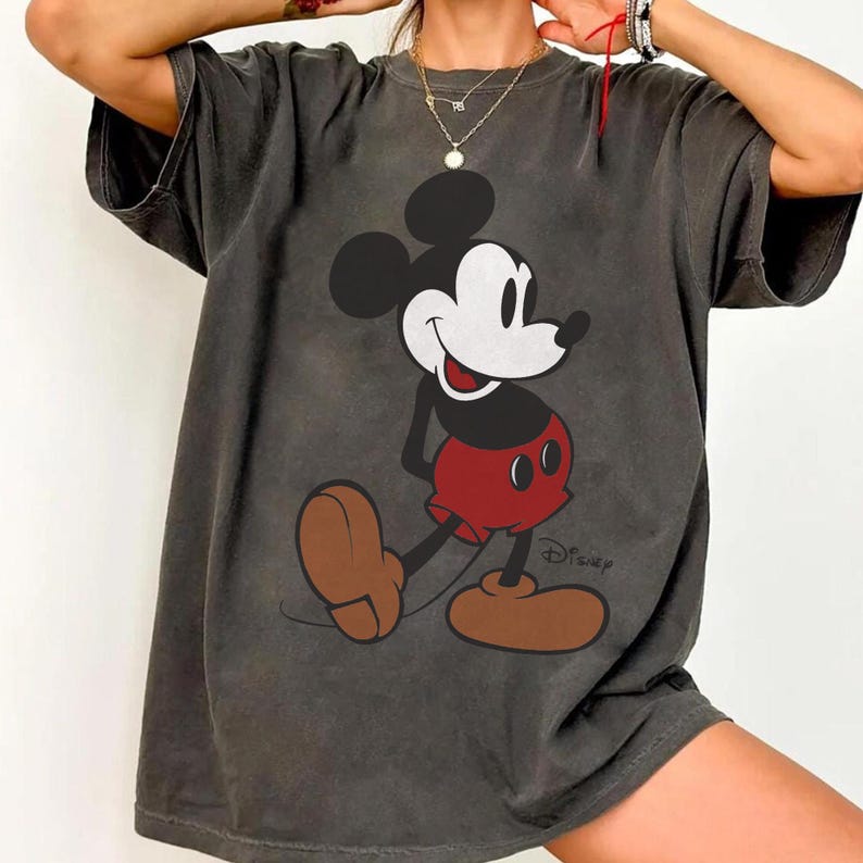 May include: Dark gray t-shirt featuring a classic Mickey Mouse graphic. The cartoon character is depicted in black, white, and red, with brown shoes. The Disney logo is printed at the bottom right. A vintage-style tee.