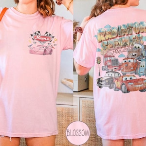 May include: A light pink t-shirt featuring a small graphic on the front chest with a race car and the text Name. The back displays a large, colorful illustration of various characters from Radiator Springs, including Lightning McQueen.