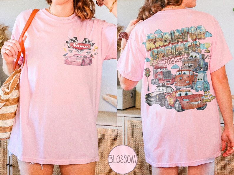 May include: A light pink t-shirt featuring a small graphic on the front chest with a race car and the text Name. The back displays a large, colorful illustration of various characters from Radiator Springs, including Lightning McQueen.
