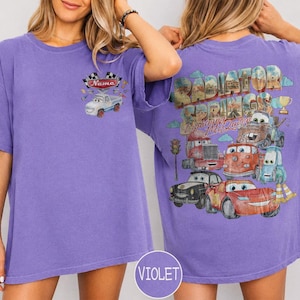 May include: A violet short-sleeve t-shirt featuring a small graphic on the front chest of a white car with checkered flags and the text Name. The back displays a large graphic of Radiator Springs with Lightning McQueen and other car characters.
