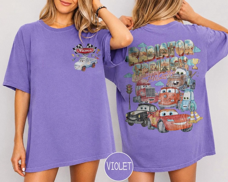 May include: A violet short-sleeve t-shirt featuring a small graphic on the front chest of a white car with checkered flags and the text Name. The back displays a large graphic of Radiator Springs with Lightning McQueen and other car characters.