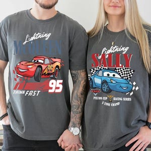 May include: Two charcoal grey t-shirts featuring Lightning McQueen and Sally Carrera from the Cars film. The Lightning McQueen shirt has the number 95 and the words "Think Fast". The Sally shirt has a chequered flag.