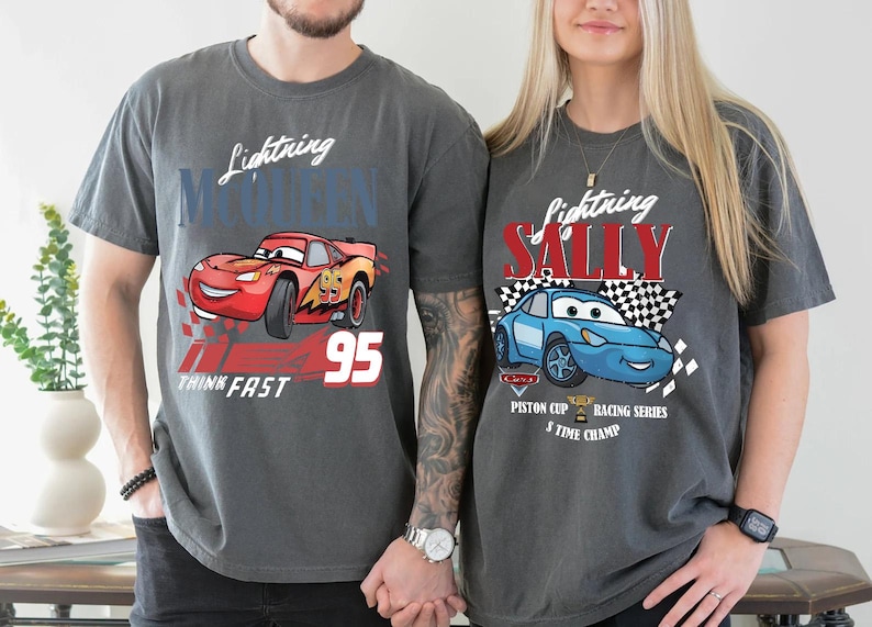 May include: Two charcoal grey t-shirts featuring Lightning McQueen and Sally Carrera from the Cars film. The Lightning McQueen shirt has the number 95 and the words "Think Fast". The Sally shirt has a chequered flag.