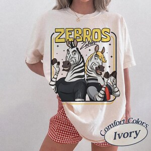 May include: Ivory-coloured t-shirt with a cartoon graphic of two zebras and the text "ZEBROS" in a yellow retro font. The zebras are wearing black and grey outfits. The shirt is a Comfort Colors brand.