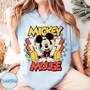 May include: Light blue t-shirt featuring a graphic of Mickey Mouse. The design includes the words "Mickey Mouse" in a retro style with yellow, red, and black colors. The shirt has short sleeves and a round neckline.