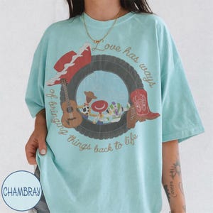 K&ouml;nnte beinhalten: Ein hellblaues T-Shirt mit einem Grafikdruck eines schwarzen Reifens, der Zeichentrickfiguren umgibt. Das Design enth&auml;lt einen roten Hut, eine braune Akustikgitarre und einen roten Cowboystiefel mit dem Text "Love has ways of bringing things back to life."