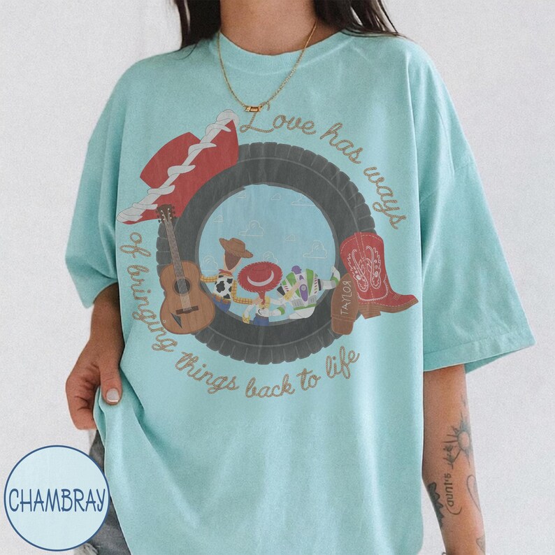 K&ouml;nnte beinhalten: Ein hellblaues T-Shirt mit einem Grafikdruck eines schwarzen Reifens, der Zeichentrickfiguren umgibt. Das Design enth&auml;lt einen roten Hut, eine braune Akustikgitarre und einen roten Cowboystiefel mit dem Text "Love has ways of bringing things back to life."