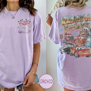 May include: A light purple t-shirt featuring a small graphic on the front chest with a race car and the text Name. The back displays a large, colorful illustration of various characters from the movie Cars with the text Radiator Springs Lightning McQueen.