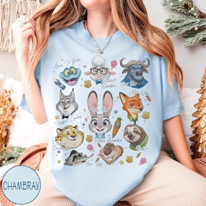 May include: Light blue t-shirt featuring cartoon illustrations of characters from a movie. The shirt includes images of a frog, sheep, buffalo, fox, rabbit, and sloth, with character names. The word "CHAMBRAY" is printed on the bottom left.