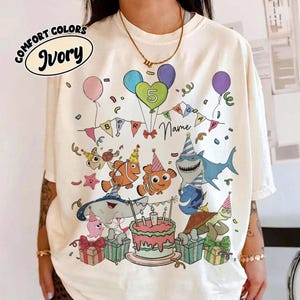 May include: An ivory t-shirt featuring a colorful graphic of sea creatures wearing party hats, including a shark, clownfish, and turtle. The design includes a birthday cake, balloons, gift boxes, confetti, and the text 5 and Name.