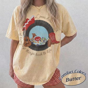 K&ouml;nnte beinhalten: Ein blassgelbes Kurzarm-T-Shirt mit einem Grafikdruck, der einen schwarzen Reifen um Zeichentrickfiguren, eine braune Akustikgitarre und einen roten Cowboystiefel zeigt. Der Text lautet Love has a way of bringing things back to life.