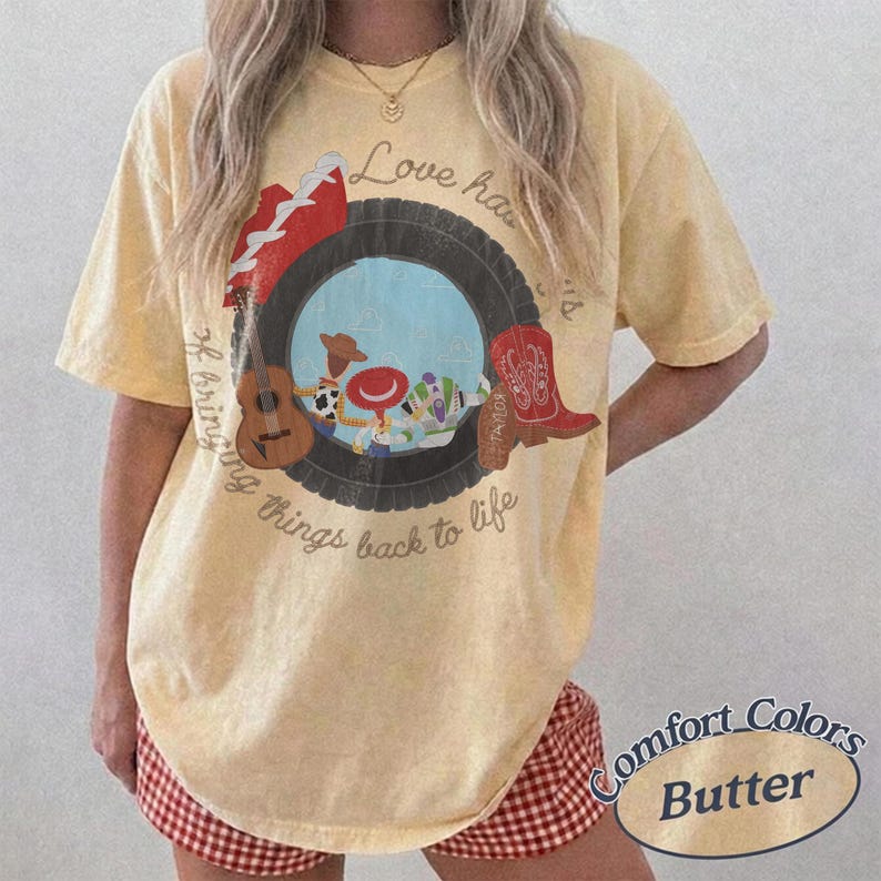 K&ouml;nnte beinhalten: Ein blassgelbes Kurzarm-T-Shirt mit einem Grafikdruck, der einen schwarzen Reifen um Zeichentrickfiguren, eine braune Akustikgitarre und einen roten Cowboystiefel zeigt. Der Text lautet Love has a way of bringing things back to life.