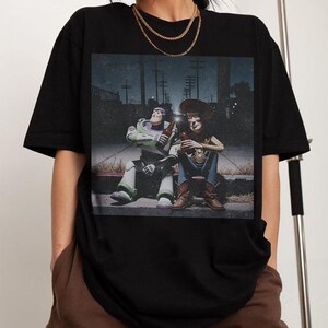 May include: Black t-shirt featuring a graphic of Buzz Lightyear and Woody from Toy Story, sitting and holding bottles. The image has a vintage, slightly faded look. The characters are depicted in a nighttime setting.