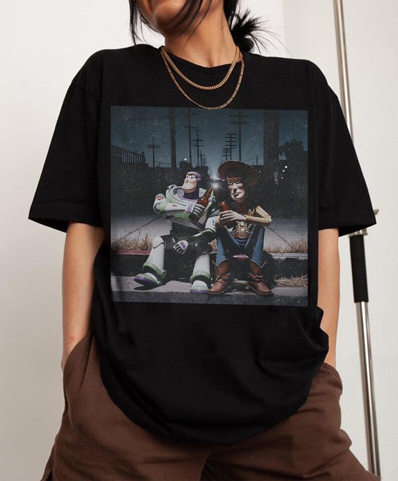 May include: Black t-shirt featuring a graphic of Buzz Lightyear and Woody from Toy Story, sitting and holding bottles. The image has a vintage, slightly faded look. The characters are depicted in a nighttime setting.