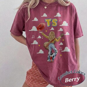 May include: A berry-colored short-sleeve t-shirt featuring a graphic of a person with long hair playing an acoustic guitar, wearing a yellow plaid shirt and cow-print bell-bottom pants. The design includes white clouds, yellow stars, and the text TS.