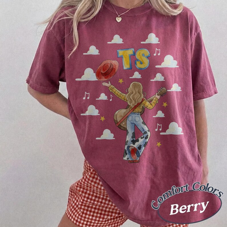 May include: A berry-colored short-sleeve t-shirt featuring a graphic of a person with long hair playing an acoustic guitar, wearing a yellow plaid shirt and cow-print bell-bottom pants. The design includes white clouds, yellow stars, and the text TS.