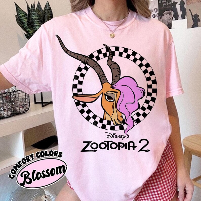 May include: A light pink t-shirt featuring a cartoon gazelle with pink hair inside a checkered circle. The shirt has the text "Disney Zootopia 2" in black. The shirt also has the text "Comfort Colors Blossom" in a circular design.