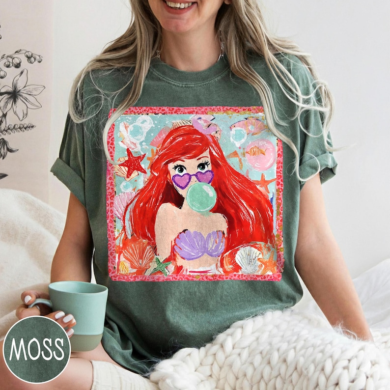 May include: Green t-shirt featuring a colorful graphic of a cartoon mermaid with red hair, heart-shaped sunglasses, and a bubble. The graphic includes seashells, starfish, and a pink border. The shirt is a relaxed fit.