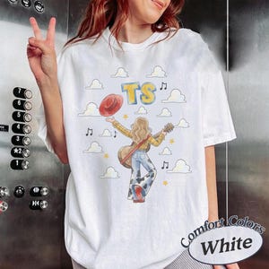 May include: A white short-sleeve t-shirt featuring a graphic of a person with long hair wearing a yellow plaid shirt and cow-print pants, holding a guitar and a red hat. The design includes blue text reading TS, white clouds, and musical notes.