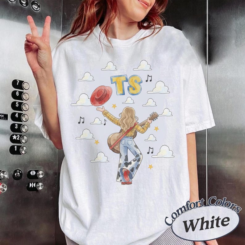 May include: A white short-sleeve t-shirt featuring a graphic of a person with long hair wearing a yellow plaid shirt and cow-print pants, holding a guitar and a red hat. The design includes blue text reading TS, white clouds, and musical notes.