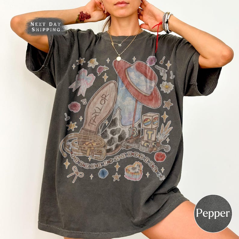 May include: A charcoal gray short-sleeve t-shirt featuring a graphic print of a cowboy boot with the name Taylor, a red hat, a belt, and a block with the letter T. The design includes small stars, flowers, and bows scattered across the front.