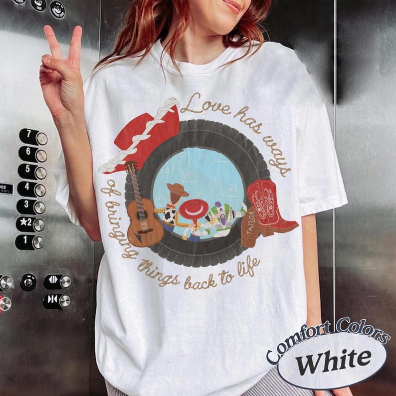 K&ouml;nnte beinhalten: Ein wei&szlig;es T-Shirt mit einer Grafik eines schwarzen Reifens, der eine blaue Himmelsszene mit Zeichentrickfiguren umrahmt. Das Design enth&auml;lt eine braune Akustikgitarre, einen roten Cowboyhut und einen roten Cowboystiefel sowie den Text Love has ways of bringing things back to life.