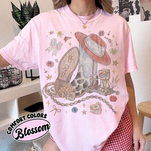 May include: A light pink t-shirt featuring a graphic print of a cowboy boot, a cowboy hat, a lasso, and alphabet blocks. The text "TAYLOR" is on the boot, "13" is on the side, and "I KNEW YOU WERE TROUBLE" is on a beaded string graphic.