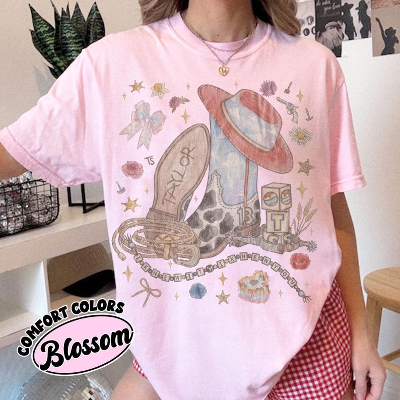 May include: A light pink t-shirt featuring a graphic print of a cowboy boot, a cowboy hat, a lasso, and alphabet blocks. The text "TAYLOR" is on the boot, "13" is on the side, and "I KNEW YOU WERE TROUBLE" is on a beaded string graphic.
