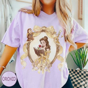 May include: A light purple t-shirt featuring a framed illustration of a woman in a yellow dress reading a book, with a red rose. The frame is adorned with a gold bow and ribbon, and the shirt has the word "ORCHID" printed on it.