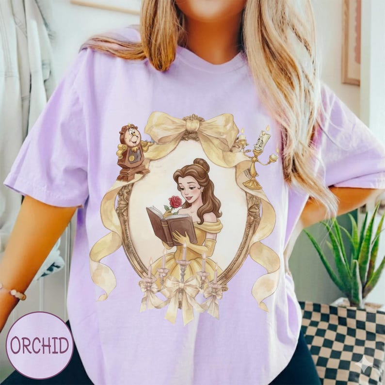 May include: A light purple t-shirt featuring a framed illustration of a woman in a yellow dress reading a book, with a red rose. The frame is adorned with a gold bow and ribbon, and the shirt has the word "ORCHID" printed on it.