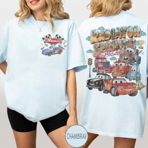 May include: A light blue t-shirt featuring a front chest graphic of a blue race car with checkered flags and the text Name. The back displays a large Radiator Springs Lightning McQueen graphic with multiple cartoon vehicles and traffic lights.