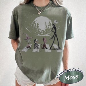 May include: A moss green short-sleeve t-shirt featuring a graphic print of characters from The Nightmare Before Christmas walking across a crosswalk. The design includes a large moon, a spooky town silhouette, and the text Comfort Colors Moss.