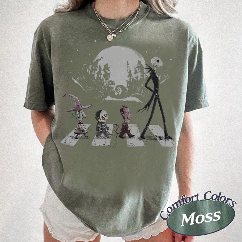 May include: A moss green short-sleeve t-shirt featuring a graphic print of characters from The Nightmare Before Christmas walking across a crosswalk. The design includes a large moon, a spooky town silhouette, and the text Comfort Colors Moss.