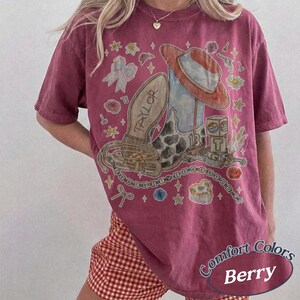 May include: A berry-colored short-sleeve t-shirt featuring a graphic print of a cowboy boot, a cowboy hat, a lasso, and a block with the letter T. The design includes the text TAYLOR and 13, surrounded by small illustrations of bows, flowers, and stars.