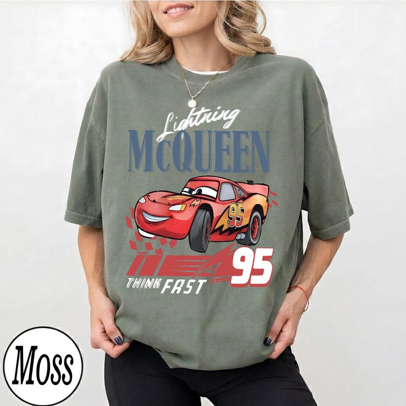 May include: Olive green t-shirt with a graphic of Lightning McQueen from the film Cars. The design includes the text "Lightning McQueen", "95", and "Think Fast". The car is red with yellow and orange accents.