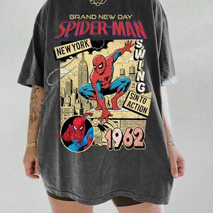 Puede incluir: Camiseta gris oscuro con un dise&ntilde;o gr&aacute;fico vintage de Spider-Man. El dise&ntilde;o incluye las palabras "BRAND NEW DAY", "SPIDER-MAN", "NEW YORK" y "1962". El superh&eacute;roe se representa en una pose din&aacute;mica sobre un fondo de paisaje urbano.